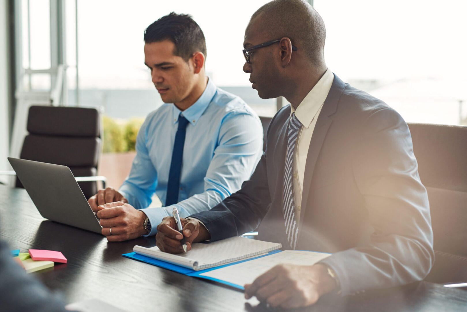 Two experienced business executives in a meeting seated at a table discussing paperwork and information on a laptop computer, one Hispanic, one African American